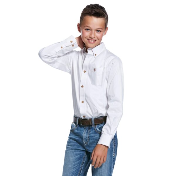 A young boy in jeans and white shirt holding his hand up to the side of his head.